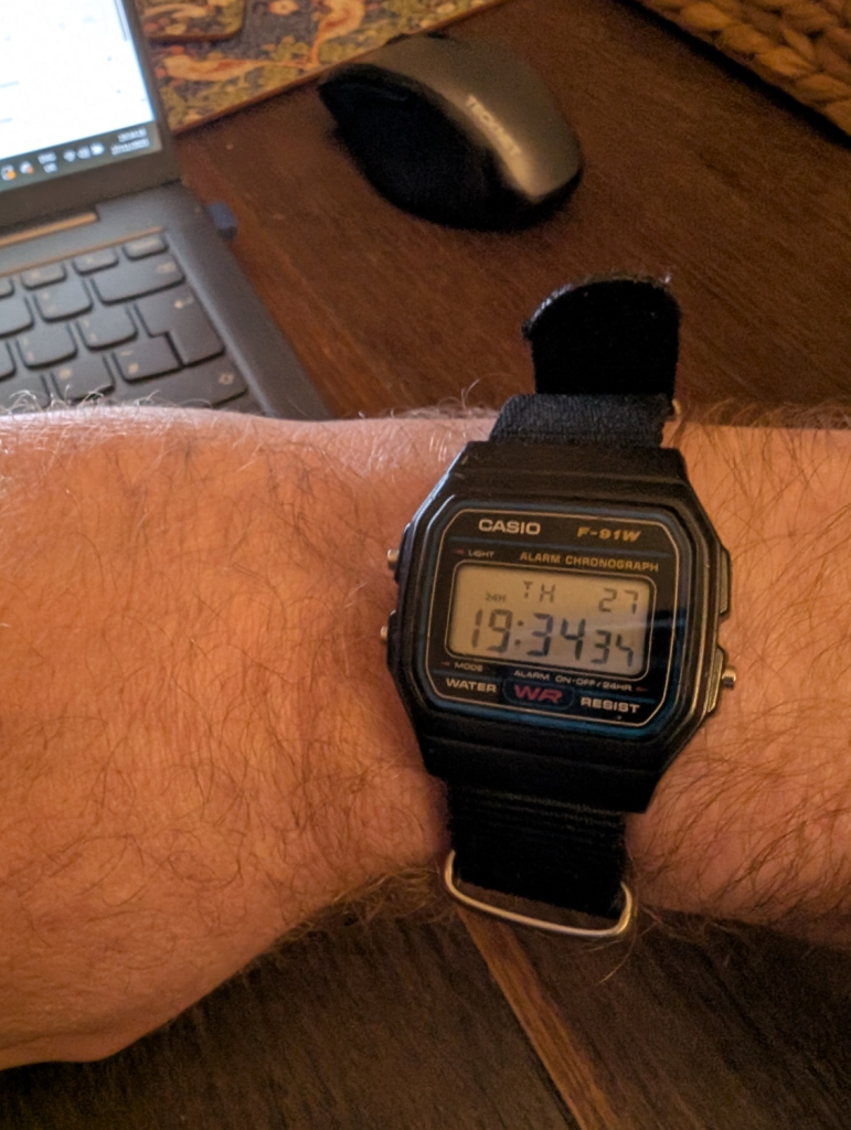 A close-up of a Casio F-91W digital watch on a wrist, fitted with a black fabric strap. The LCD display reads "TH 27" and the time is 19:34. In the background, a laptop keyboard and wireless mouse sit on a wooden desk.