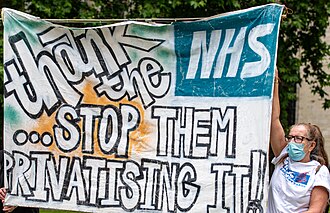 A protester wearing a face mask holds up a large hand-painted banner outside the UK Parliament. The banner reads "Thank the NHS... STOP THEM PRIVATISING IT!" in large black and white lettering.