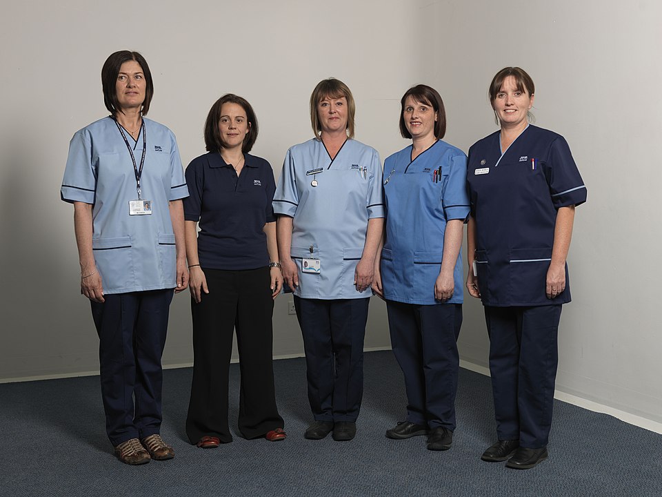 Five NHS Scotland healthcare workers standing in a row against a white background, wearing various blue and navy uniforms representing different clinical roles.