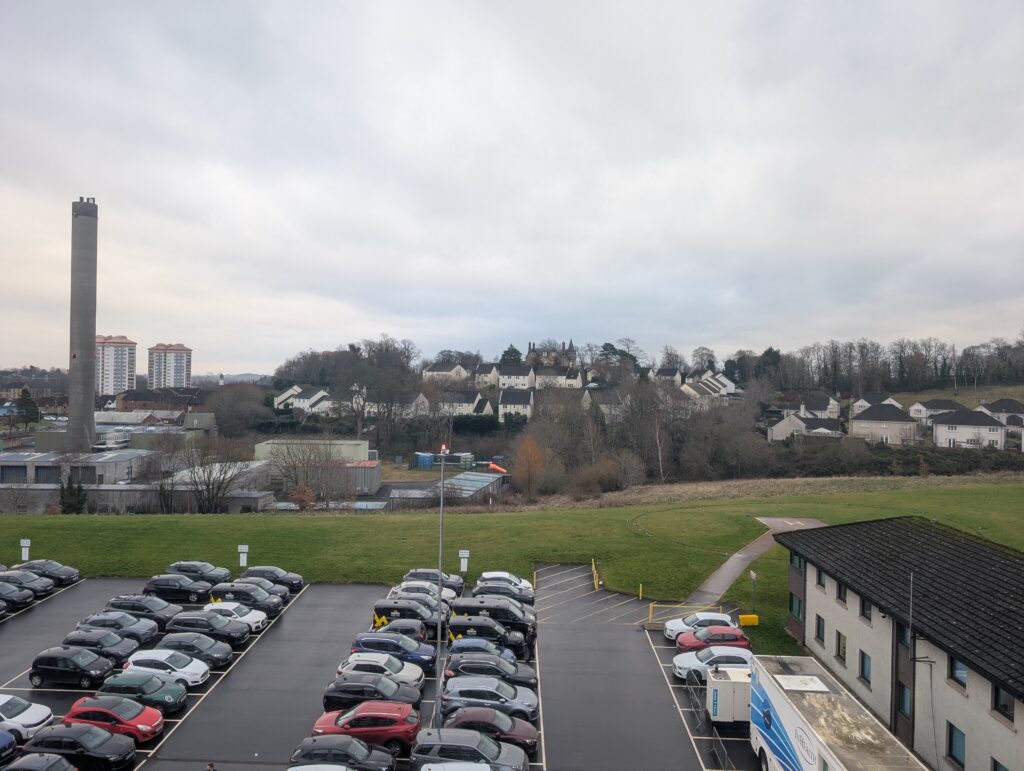 A view from a high hospital window looking out over Paisley on a cloudy day. In the foreground is a large car park filled with vehicles. To the left stands a tall, thin concrete industrial chimney. In the background, a mix of modern suburban housing and high-rise flats sits in front of rolling green hills. A grassy area in the middle ground contains a helicopter landing pad.