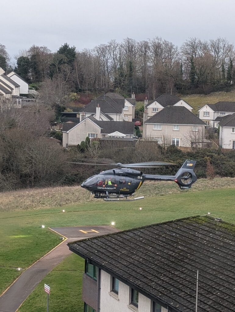 A dark grey air ambulance helicopter hovering just above a grass landing pad next to a hospital building. In the background are suburban houses and bare winter trees under a grey sky.