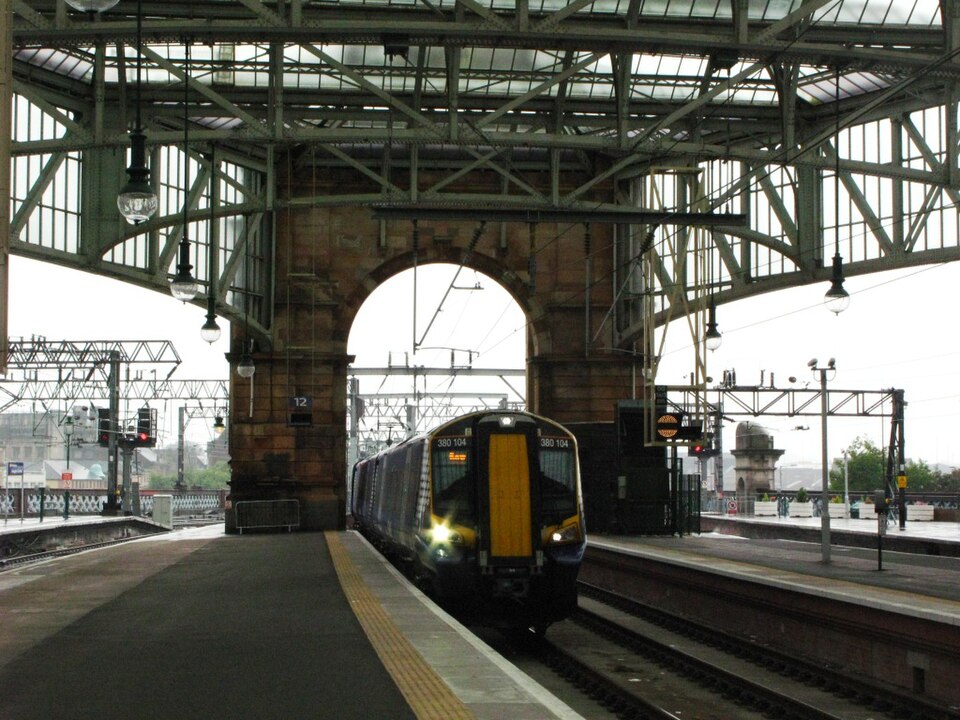 A ScotRail Class 380 train arrives at platform 12 of Glasgow Central station, framed by a Victorian stone arch and the station's ornate iron and glass roof.

