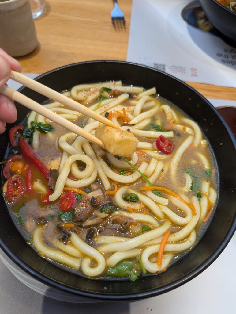 A black bowl of Kare Burosu, thick udon noodles in a golden curry broth with mushrooms, red chillies, shredded carrot and coriander, with a piece of silken tofu held between chopsticks