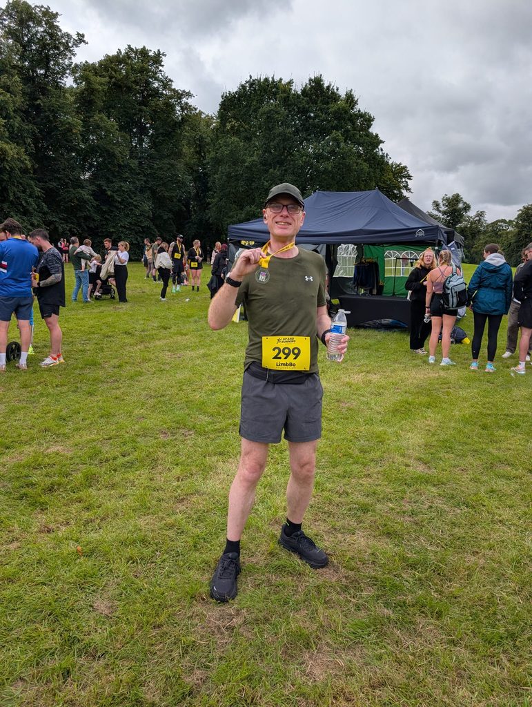 Douglas standing on grass near the finish area, smiling and holding up his medal, with his race number still pinned on and a water bottle in his other hand.