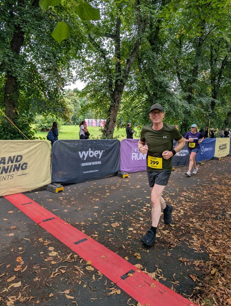 Douglas running along a tree-lined park path, smiling, with race barriers and other runners visible behind him.