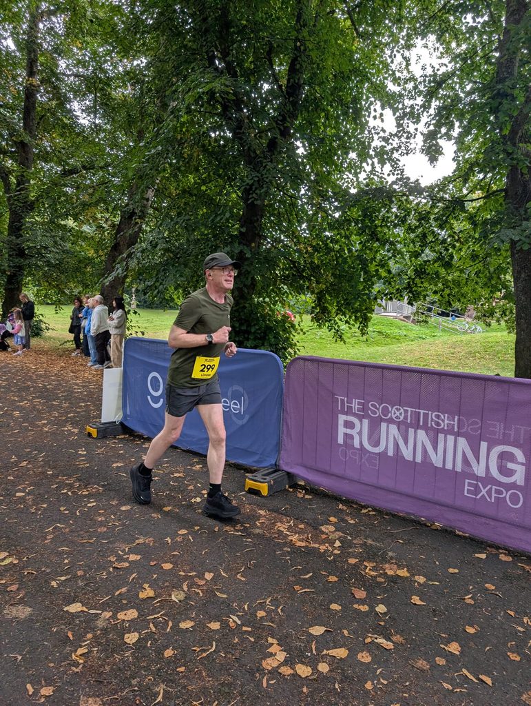Douglas running with a strained, focused expression along a leaf-covered path lined with event barriers.