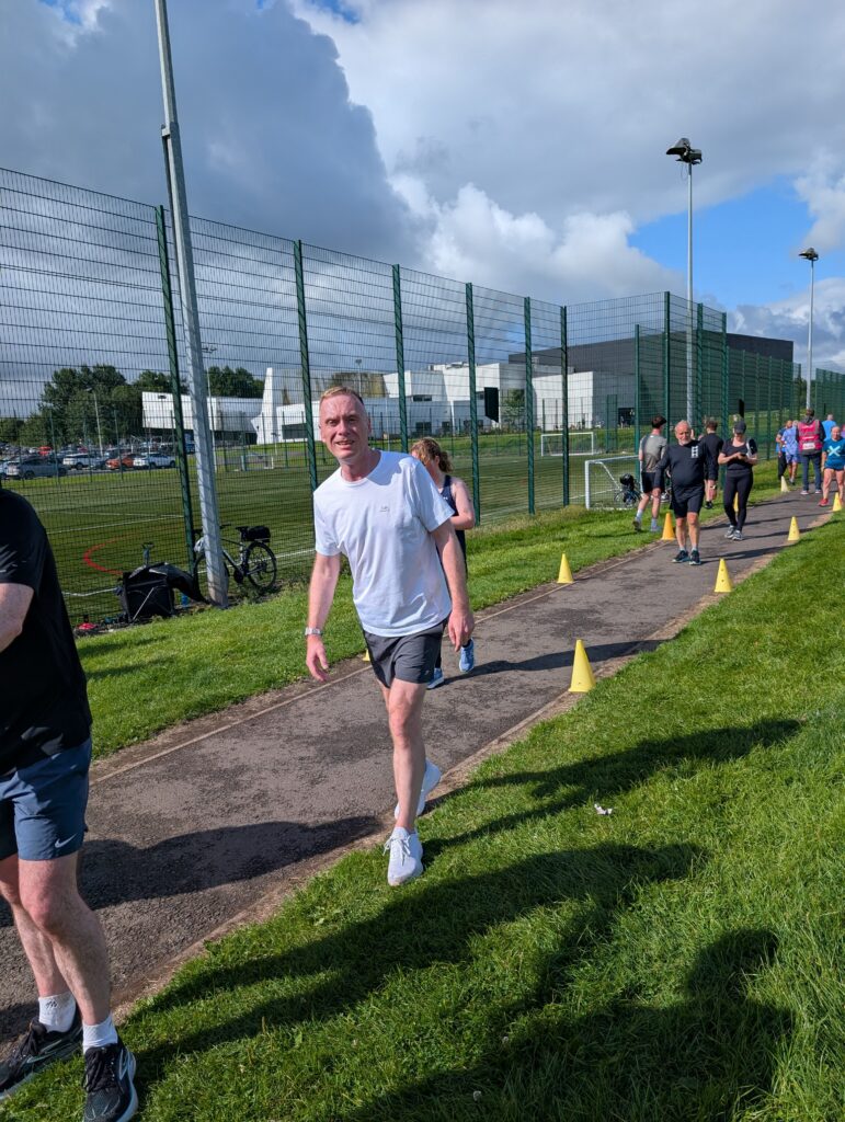 A man in a white t-shirt and dark shorts running along a tarmac path beside a sports pitch fence, other parkrunners visible behind him, under a partly cloudy sky.