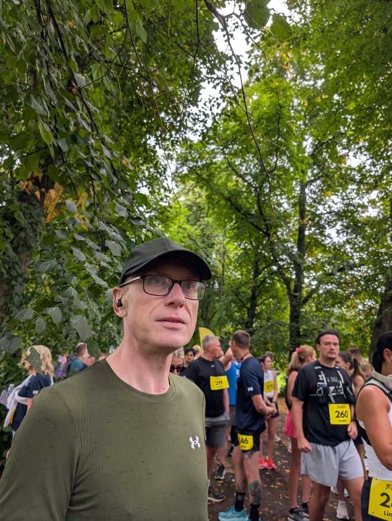 Close-up of Douglas standing among other runners under trees at the start area, wearing a cap and glasses, looking slightly tense.