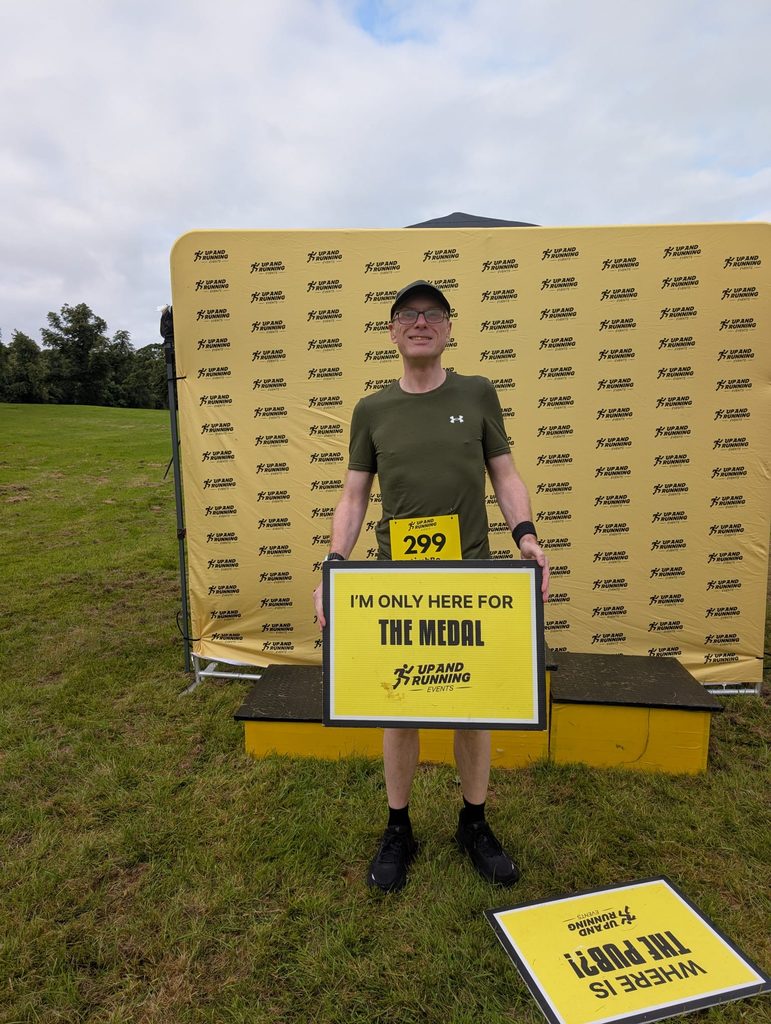 Douglas standing in front of an Up and Running Events photo backdrop, holding a printed sign reading 'I'm only here for the medal'.