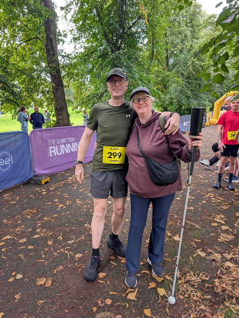 Douglas in his running kit and race number with his arm around Stephanie, who is wearing a hoodie and holding a long cane, on the path near the start line.