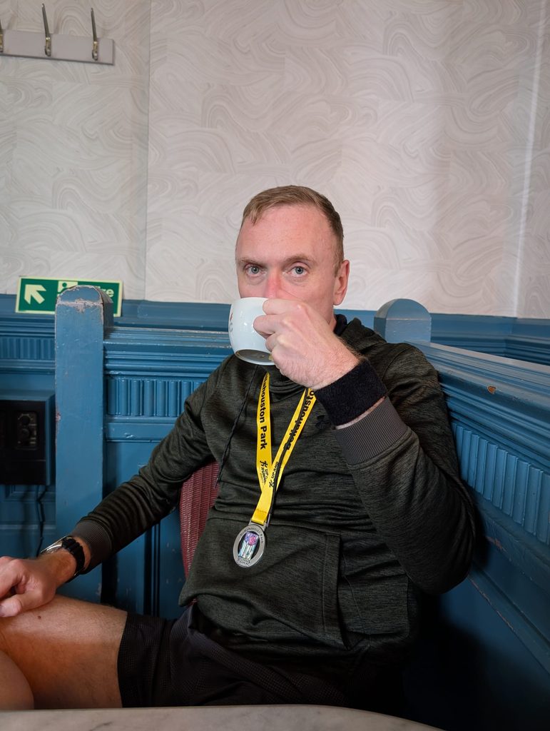 Douglas sitting in a café booth drinking from a cup, still wearing his finisher's medal over a hoodie.